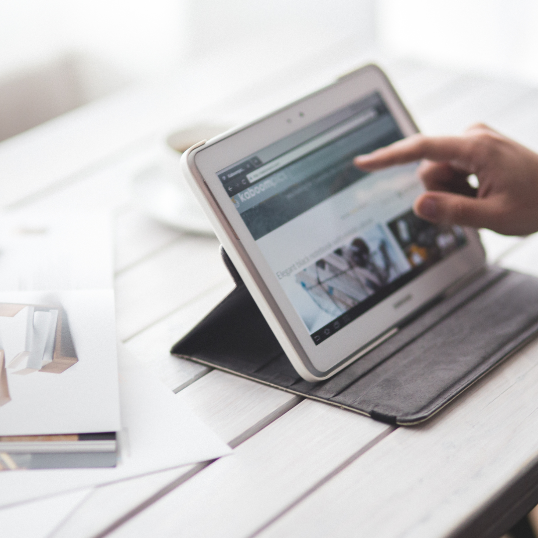 يد تستخدم جهازًا لوحيًا لزيارة موقع إلكتروني على طاولة عمل بيضاء مع أوراق وتصاميم مطبوعة، في إشارة إلى متابعة نتائج الحملات التي تديرها أفضل شركات التسويق الإلكتروني.
